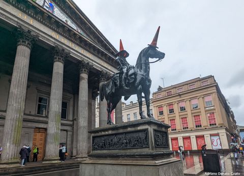 Em frente a um prédio com grandes e imponentes colunas cinza, vemos uma escultura equestre preta. Cones laranja de trânsito estão em cima da cabeça do cavalo e do cavaleiro.