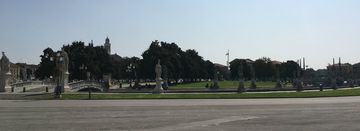 Uma praça oval logo ao centro da imagem, toda rodeada de estátuas, cada uma sobre uma coluna. Há também uma ponte conectando a borda da praça com seu interior, passando por cima de um pequeno canal. Muitas árvores no centro, sob um céu bem claro.