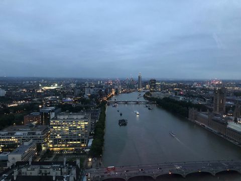 Em um início de noite bem nublada, vê-se o rio Tâmisa ao centro com edifícios de diferentes tamanhos em ambas as margens, a maioria já com suas luzes acesas.