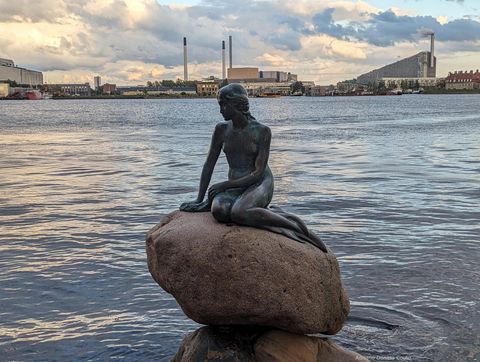 'Em um grande canal, vemos em primeiro plano a escultura de uma sereia sentada sobre uma pedra, observando o horizonte. Ao fundo, prédios de diferentes alturas com longas chaminés.'