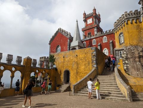 'A partir de uma área de piso de concreto, vemos algumas torres e espirais em tons de vermelho e amarelo. É um palácio. À esquerda, temos um mirante; á direita, uma escada que leva aos níveis superiores da construção.'