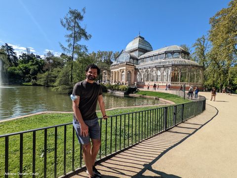 Um rapaz de camiseta marrom e bermuda azul posa em frente a um lago com várias árvores em volta. Ao fundo, um palácio feito de vidro e metal.