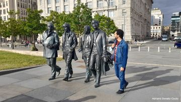 No plano de fundo, uma via asfaltada e um prédio antigo. Em primeiro plano, uma estátua de bronze de 4 rapazes caminhando enquanto conversam. Ao lado, um rapaz moreno de cabelos encaracolados, vestindo jaqueta e calça jeans.