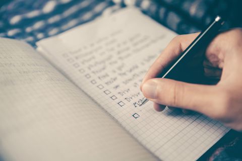 Uma mão segura uma caneta preta clássica sobre um caderno de folhas quadriculadas, com uma lista de itens escrita em uma das folhas.