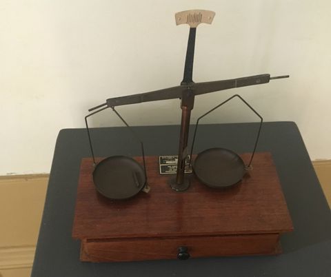 Balança de dois pratos em metal escuro, com base em madeira. O objeto está sobre uma superfície cinza escuro.