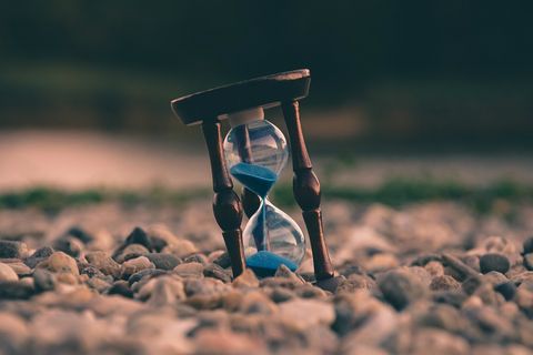 Em uma área cheia de pedregulhos, uma ampulheta está apoiada de forma inclinada. Por dentro, uma areia azul escorre para o compartimento inferior.