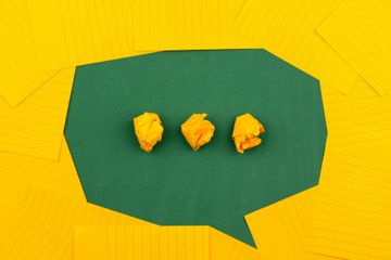 Folhas de papel amarelas distribuías sobre uma superfície verde foram um balão de conversa, como os utilizados em histórias em quadrinho. No centro, reticências amarelas também de bolinhas de papel.