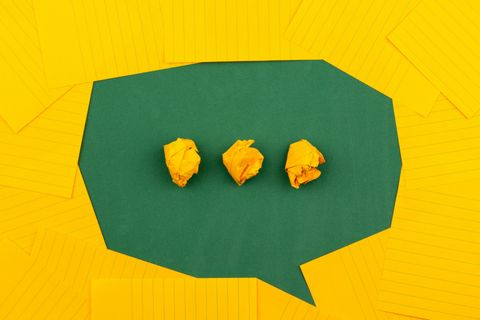 Folhas de papel amarelas distribuías sobre uma superfície verde foram um balão de conversa, como os utilizados em histórias em quadrinho. No centro, reticências amarelas também de bolinhas de papel.