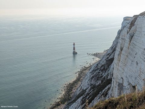 'À direita, um penhasco de paredes brancas; à esquerda, em nível inferior, um farol no meio do mar, próximo da costa.'