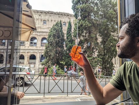 Um rapaz moreno de cabelo encaracolado está vestindo camiseta verde. Ele (em perfil) levanta uma taça com drink laranja. Ao fundo, uma arena romana antiga.