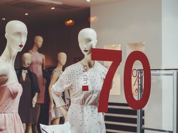 No vidro de uma vitrine, um adesivo enorme indica 70% de desconto. Logo atrás, alguns manequins femininos com vestidos de cor mais neutra. Eles não possuem o olho marcado, mas os lábios são vermelhos.