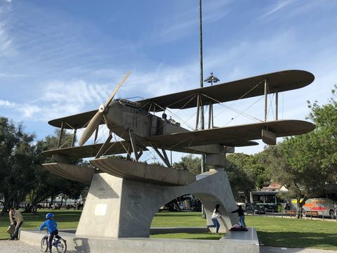Monumento com um avião de metal em tom próximo do bronze escurecido. Ele possui um par de hélices no bico e duas estruturas que lembram boias na parte inferior. O avião está sobre uma estrutura de concreto, em uma região arborizada.