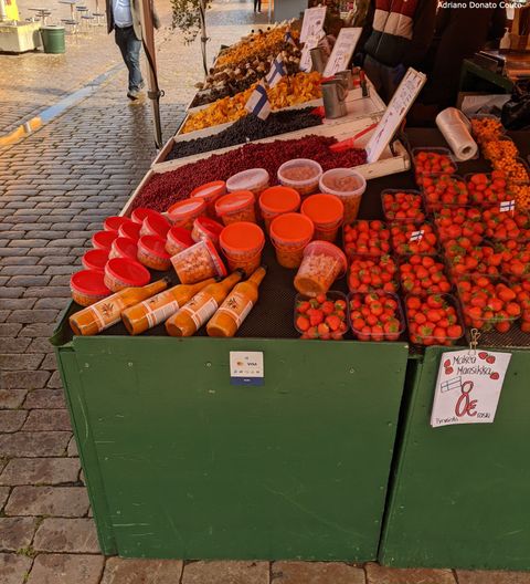 "Em uma praça coberta de paralelepípedos, uma barraca verde exibe frutos, molhos e cogumelos de diferentes tons, majoritariamente de vermelho e amarelo."