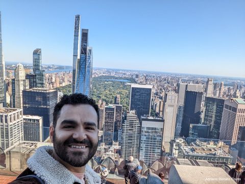 Um rapaz sorri em primeiro plano, no topo de um prédio. Em segundo plano, uma série de prédios e uma faixa verde (Central Park).