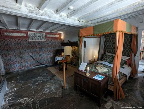 Vemos o interior de um quarto em um imóvel antigo. O teto tem vigas de madeira; o piso, todo de placas irregulares de pedra. Há uma cama com proteção superior ricamente ornada, além de uma mesinha de madeira.