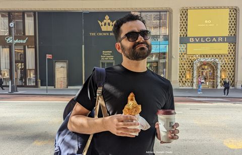 Rapaz moreno, com camiseta preta e óculos escuros, mantém um semblante sério enquanto segura um croissant e um café. Ao fundo, fachada de lojas.