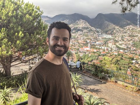 "Rapaz moreno (eu) com camiseta marrom em primeiro plano. Ao fundo, uma colina repleta de casas e uma cadeia de montanhas, com o topo coberto por nuvens."