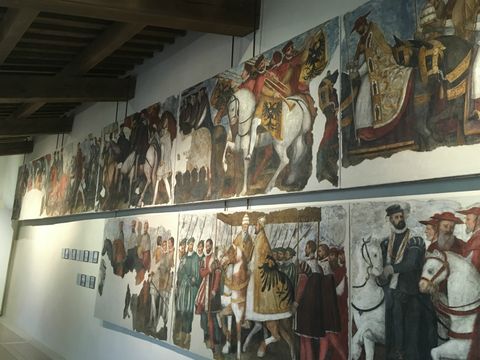 Parede branca com uma série de fragmentos de afrescos. Nas pinturas, são retratados cavaleiros e líderes montados em seus cavalos, em uma espécie de procissão.