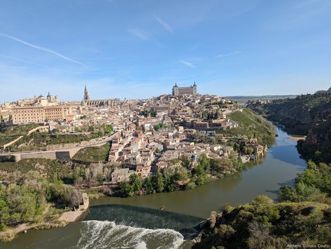 Em primeiro plano, um rio ladeado por vetação. Logo atrás, cercada pelo rio, uma colina repleta de construções medievais, incluindo algumas torres de igreja e uma fortaleza militar.
