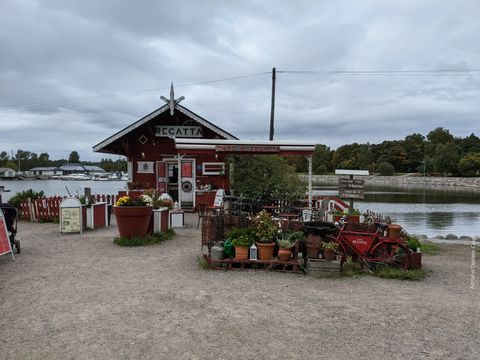 "Em primeiro plano, um conjunto de vasos com flores e uma bicicleta vermelha. Logo atrás, uma pequena casinha vermelha, local de atendimento do Café Regatta. Ao fundo, uma espécie de marina com alguns barcos."