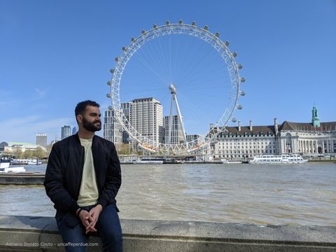 Rapaz sentado às margens do rio Tâmisa, com a roda-gigante London Eye ao fundo.