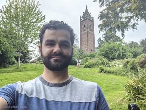 Selfie de um rapaz moreno de cabelo encaracolado, vestindo camiseta azul e cinza. Ao fundo, uma torre antiga (Cabot Tower) e área verde com árvores (Brandon Hill).
