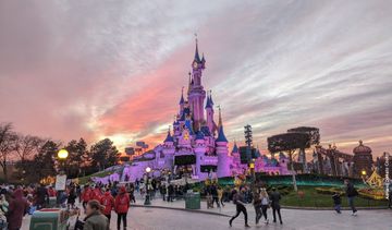 Sob um céu de fim de tarde, temos um castelo rosa com torres de telhado azul. Em frente, uma rua com muitas pessoas caminhando.