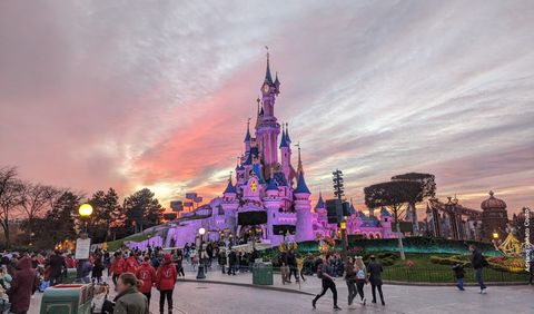 Sob um céu de fim de tarde, temos um castelo rosa com torres de telhado azul. Em frente, uma rua com muitas pessoas caminhando.