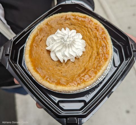Em um recipiente preto de plástico, uma forminha de alumínio com uma torta alaranjada de abóbora, que tem um pouco de chantilly no topo.