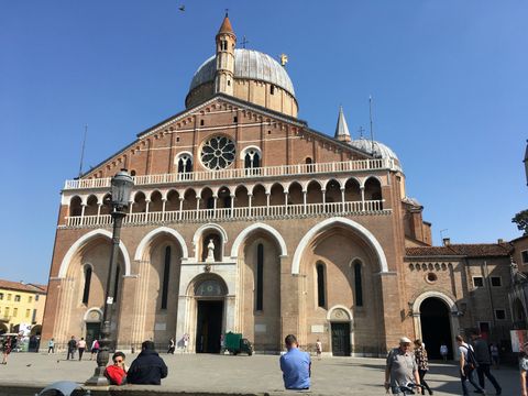 Basilica di Sant'Antonio di Padova. Enorme basílica em tom avermelhado como tijolo, com diversos pilares no topo. Há uma grande cúpula central. Logo em frente, algumas pessoas circulam pelos arredores.