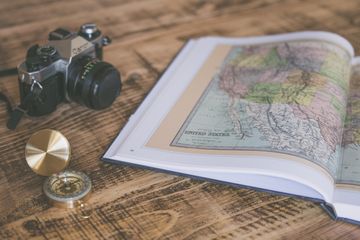 Superfície em madeira, com um livro aberto por cima, exibindo um mapa. Ao seu lado, uma bússola e uma câmera antiga.