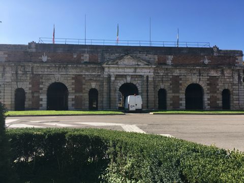Um arbusto retangular e uma rua de asfalto fazem frente a uma muralha de pedra, sob um céu azul sem nuvens. Há várias portas. Sob uma delas, há uma van branca estacionada.