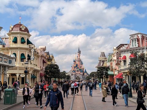 Uma rua ladeada por lojas coloridas e recheada de transeuntes leva o visualizador a um castelo rosado.