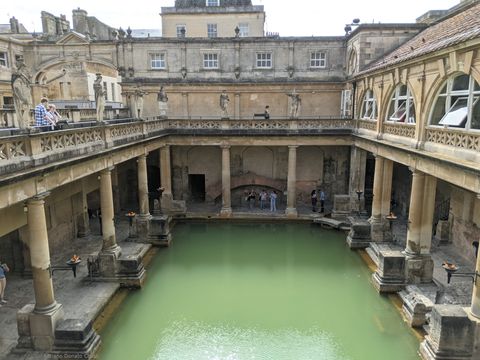 Termas romanas, com enorme piscina esverdeada ao centro, ladeada por passagens cobertas, que são sustentadas por grossas colunas de pedra. O ângulo da vista é a partir de cima.