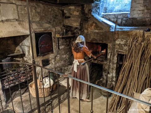 Uma espécie de cozinha medieval, toda em parede de pedra, com um fogão à lenha à esquerda. Ao centro, uma figura representando uma mulher, com blusa laranja e saia longa marrom, toda suja de terra. Ela opera um forno de pedra.