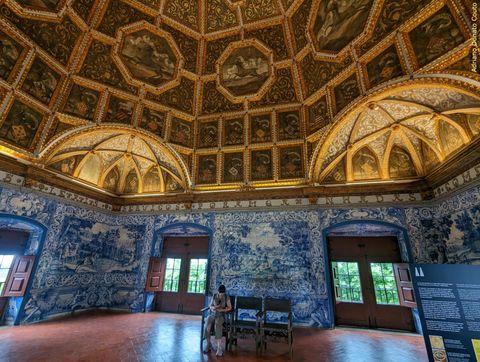 'Vemos parte de uma sala de piso de azulejos vermelhos, com paredes cobertas em azulejos azuis e brancos. O teto é coberto de molduras e pinturas, em estrutura que parece ser de madeira.'