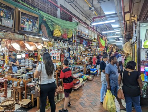 Um corredor lotado de pessoas, com muitos produtos expostos, entre eles caldeirões, peças de artesanato e itens de tecidos de diversas cores.