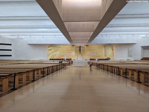 Área interior de uma basílica moderna, em tons claros. Vê-se duas fileiras de bancos ladeando um amplo corredor. Ao fundo, um painel dourado e um crucifixo logo em frente.