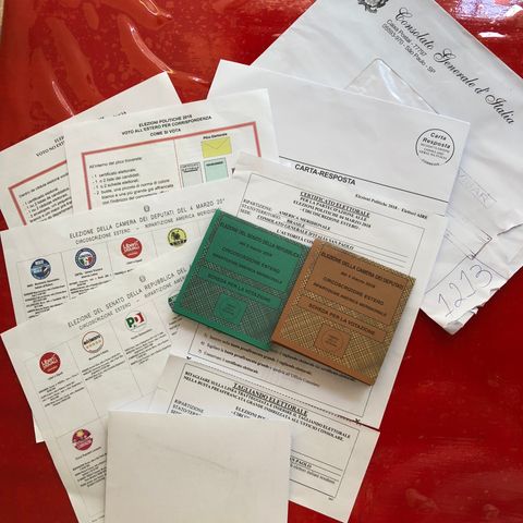 SObre uma superfície vermelha, vários documentos e envelopes em papel branco, além de duas cédulas de votação, sendo uma verde e uma marrom.