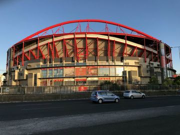 Em primeiro plano, uma via asfaltada com dois carros em tons metalizados. Logo atrás, um estádio de futebol com armação metálica vermelha e estrutura de concreto, com alguns banners colados na fachada.