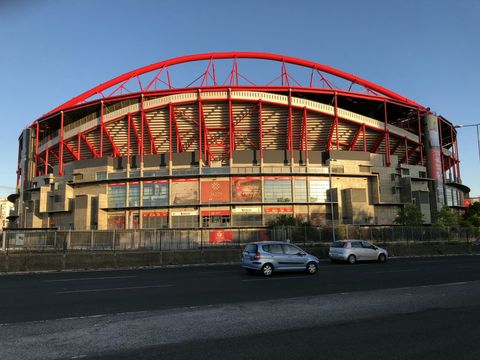 Em primeiro plano, uma via asfaltada com dois carros em tons metalizados. Logo atrás, um estádio de futebol com armação metálica vermelha e estrutura de concreto, com alguns banners colados na fachada.
