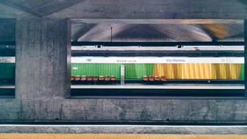 Através de um vão aberto em uma parede cinza de concreto, vê-se parte da plataforma de uma estação de metrô. Acima, uma faixa branca com o nome da estação "Vila Mariana". Logo abaixo, uma sequência de faixas em verde e amarelo na parede, com uma fileira de bancos de encosto marrom.