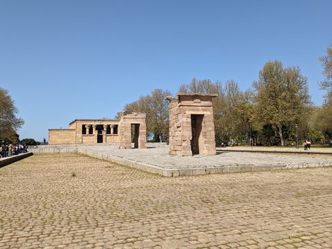Uma área pavimentada com pedras em formato de paralelepípedo rodeia uma plataforma com dois portais de pedra e, ao fundo, um templo cúbico com quatro colunas na frente. Céu azul.