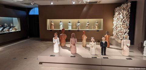 Em uma sala com paredes de tons terrosos, vemos uma série de esculturas de barro, retratando noivas de vários tons de pele e um noivo. São peças artesanais.