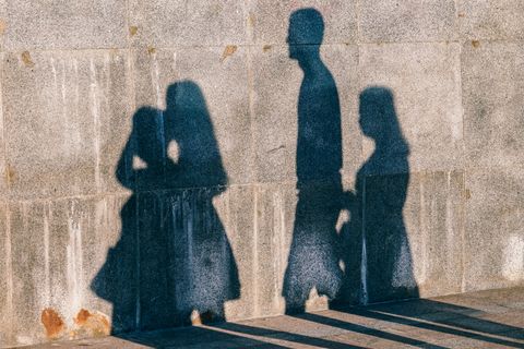 Sobre uma parede de cimento, há a projeção da sombra de quatro pessoas em perfil, entre homens e mulheres.