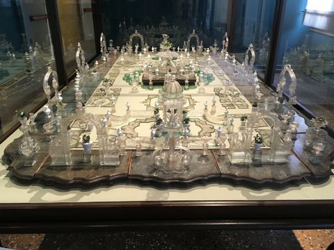 Uma superfícei em madeira, com o centro claro e cheio de desenhos, simulando uma praça. Por toda a área, colunas, arcos e escultura em vidro complementam a paisagem. Uma espécie de maquete.
