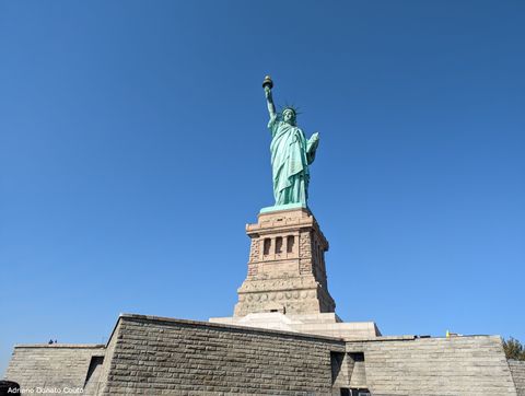 Sob céu azul sem nuvens, vemos uma base de tijolos cinza, com uma estátua azulada de bronce no topo. A escultura representa uma mulher com uma túnica. Em seu braço esquerdo, ela segura uma tábua com inscritos; na mão esquerda (com braço erguido), segura uma tocha.