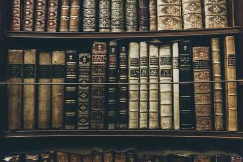 Parte de uma estante repleta de livros antigos. A maioria deles carrega tons de marrom nas capas, com detalhes em dourado.