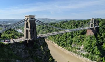 Ponte suspensa sobre um profundo leito de rio, o rio Avon. Nas margens, extensa área de mata.