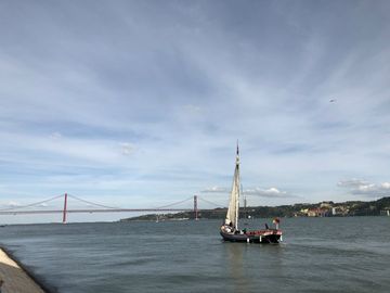 Uma pequena embarcação à vela navega pelo leito amplo do rio Tejo, que lembra o mar. Ao fundo, uma fonte suspensa de metal, com um vão amplo entre suas pilastras.
